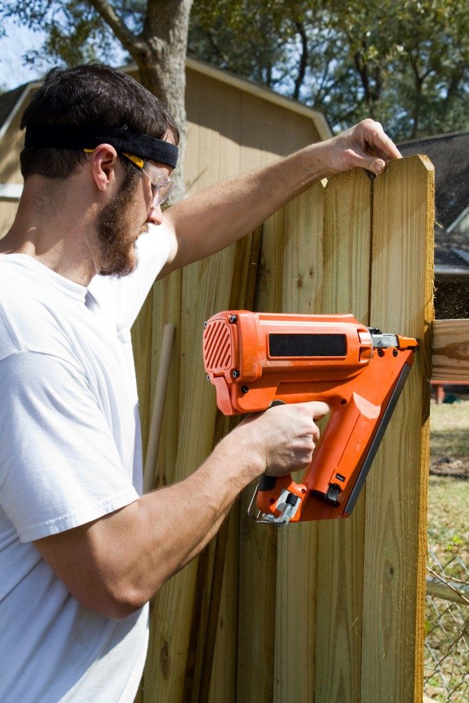 Man wearing safety glasses uses a portable nail gun to attach wood pickets to the rail as he builds a privacy fence in the backyard.