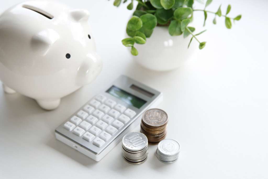 a white piggy bank, calculator, some coins and a white flower vase laid on a table